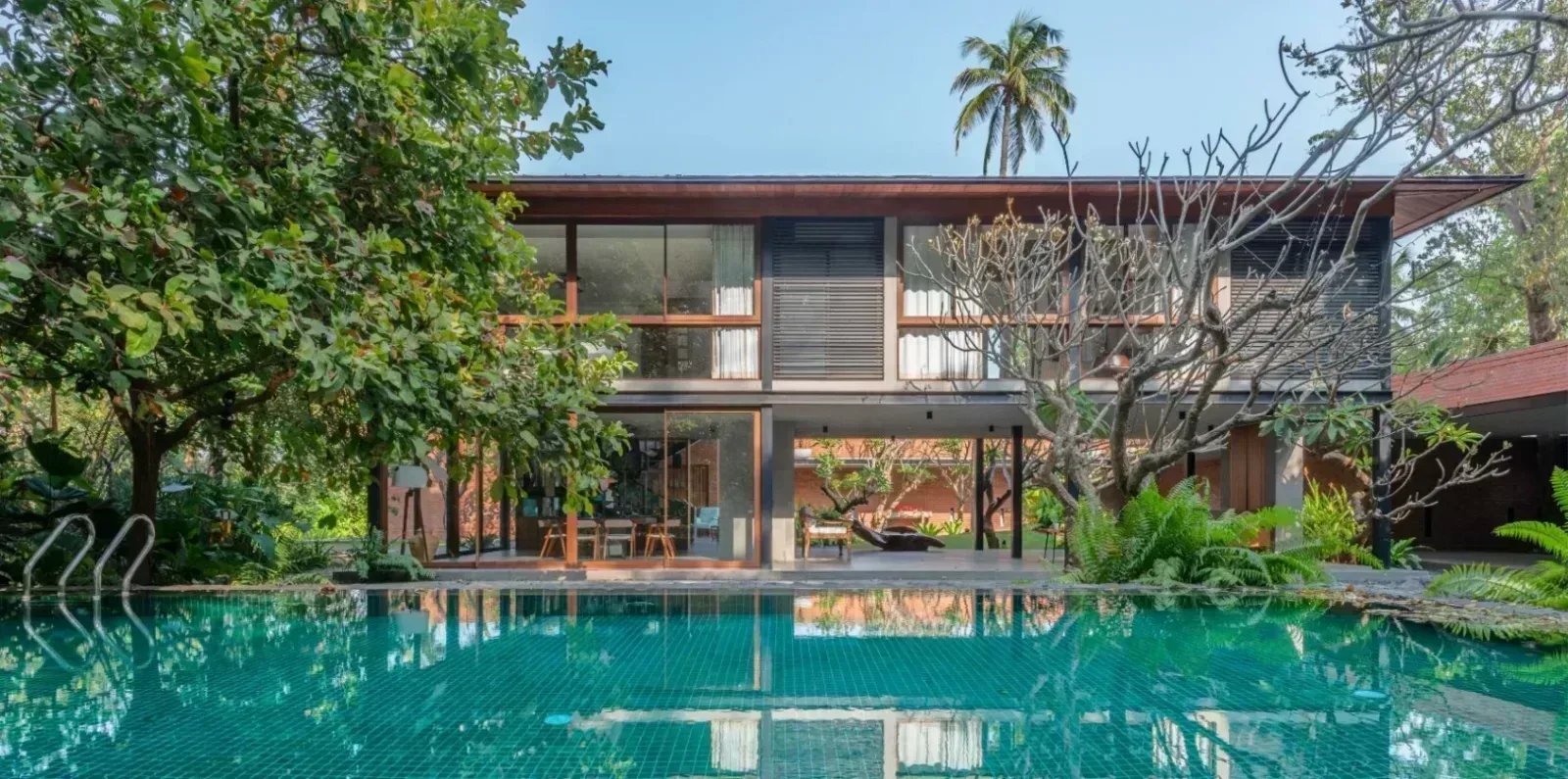 Modern luxury home with a natural exposed brick facade, large glass windows, lush landscaping, and a swimming pool.