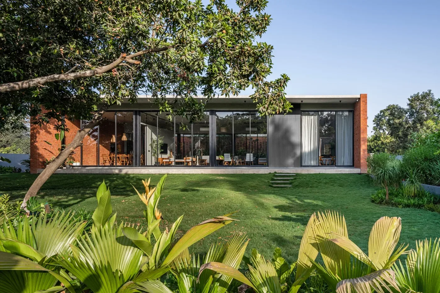 Modern home with exposed red brick walls, large glass windows and lush green landscaping in a contemporary architectural design.